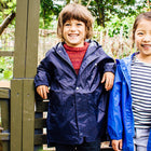 A smiling child in a Originals Waterproof Jacket Navy stands by a wooden fence, with another child partially visible to the right.